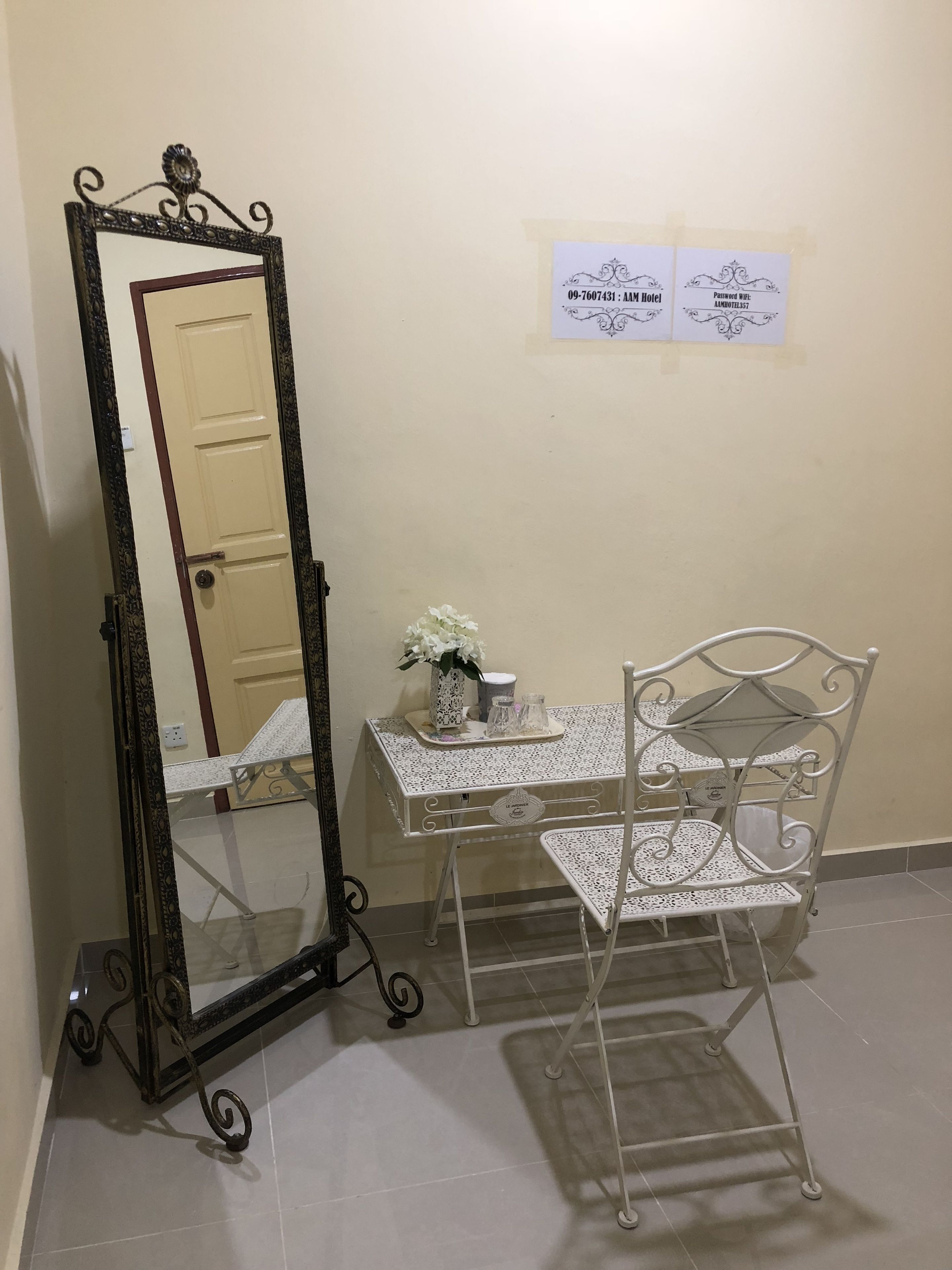 A room with a white metal desk, a white chair, and a large mirror with a decorative frame. There are two signs on the wall, one with a hotel name and another with a password.