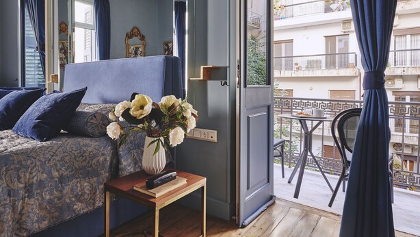A bedroom with a bed, a bedside table with a vase of flowers, and a view of the balcony. - Bohemian Suites Athens - Maison Hotel Adults Only (Athens)