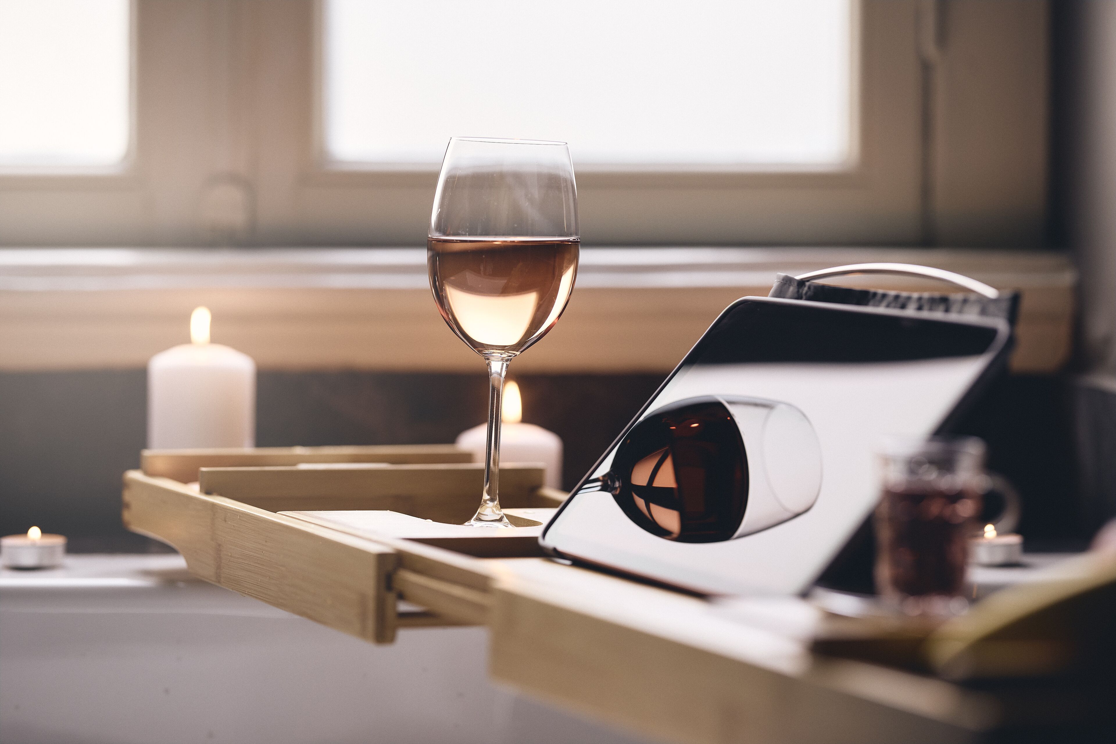 Una copa de vino blanco sobre una bandeja con un cuaderno, gafas de sol, una vela y una taza de té en una mesa.