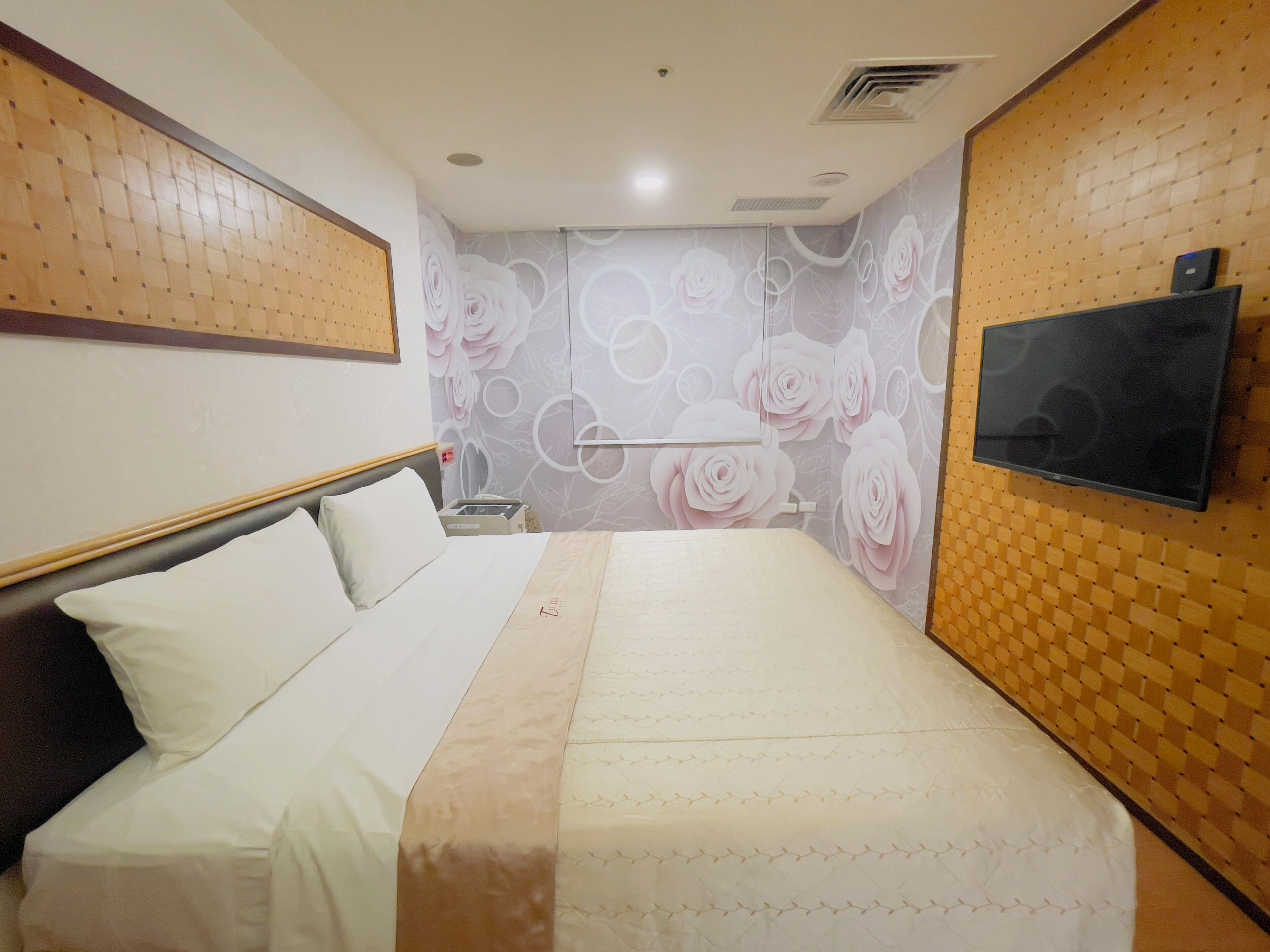 Een hotelkamer met een bed, een aan de muur bevestigde televisie en een decoratieve wand met bloemenmotieven.