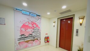 Um saguão de hotel com um mural tradicional japonês, um extintor de incêndio, um elevador e um balcão de recepção.