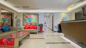 Um saguão de hotel com uma mesa de recepção, um sofá com almofadas vermelhas, uma mesa de centro e um grande quadro abstrato na parede.