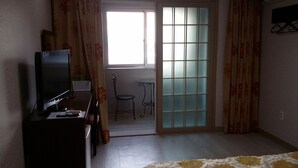 Una habitación pequeña con una televisión sobre un mueble bajo, una mesita y una silla. Se aprecia una ventana con paneles de vidrio esmerilado y cortinas.