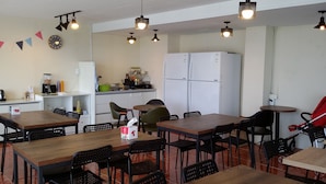 Comedor con mesas y sillas de madera, zona de cocina con refrigerador y pileta, y un reloj en la pared.
