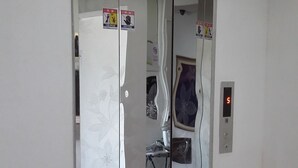 Un ascensor con puerta de vidrio reflectante, un letrero de salida de emergencia en rojo y un extintor en el suelo.