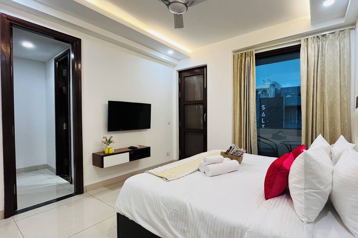 Habitación de hotel moderna con una cama, un televisor de pantalla plana, un pequeño estante con adornos y una puerta corrediza de vidrio que da a un balcón.