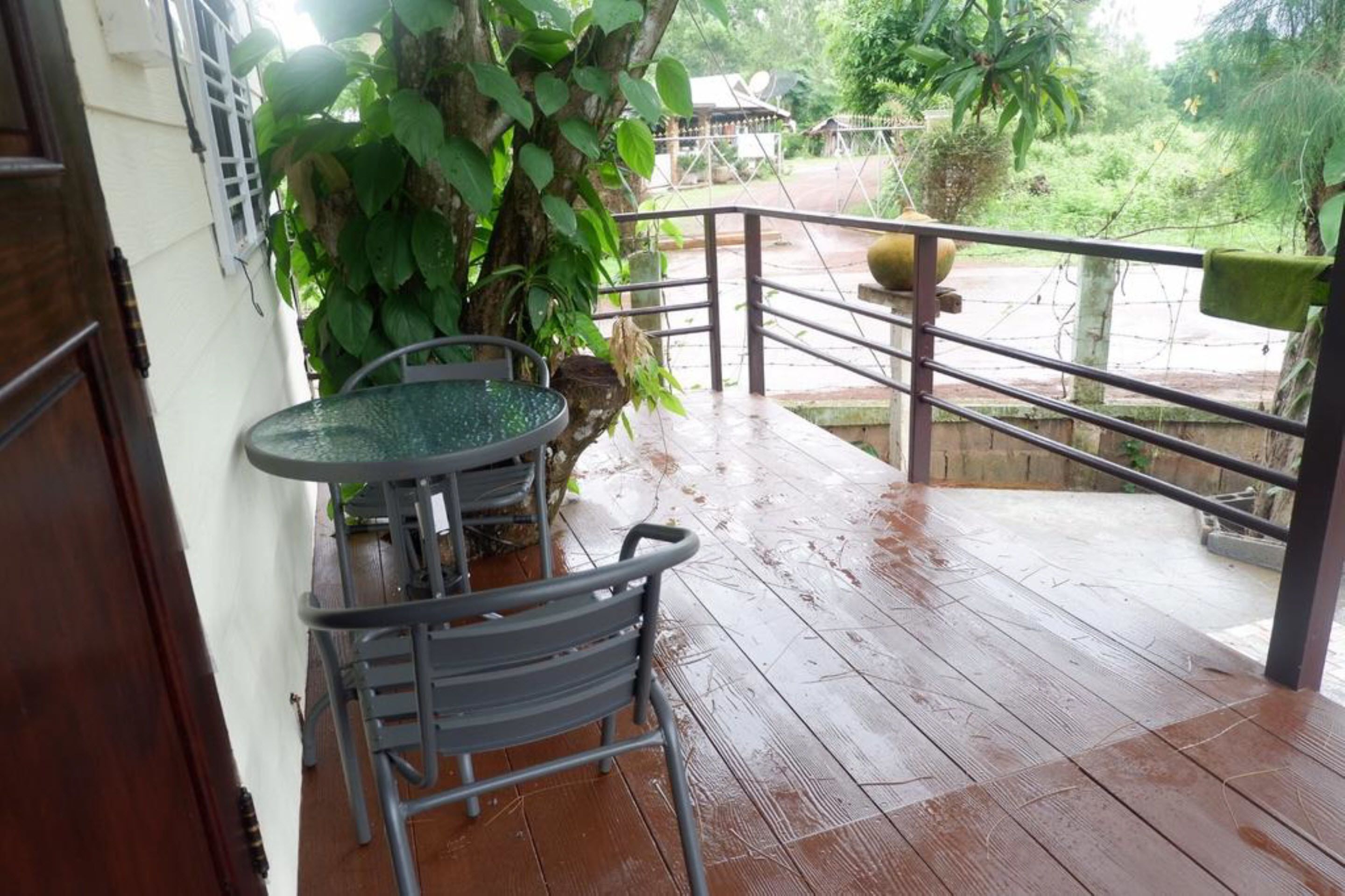 Une terrasse en bois, humide, avec une table et des chaises en métal, entourée de verdure et d’une rambarde en métal.