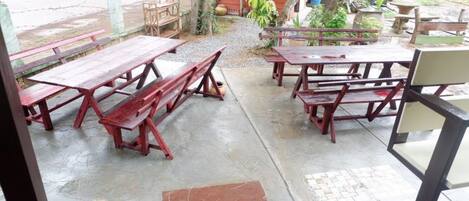 Un espace extérieur aménagé avec plusieurs tables et bancs de pique-nique en bois, un sol en béton et une vue sur une route et la verdure en arrière-plan.