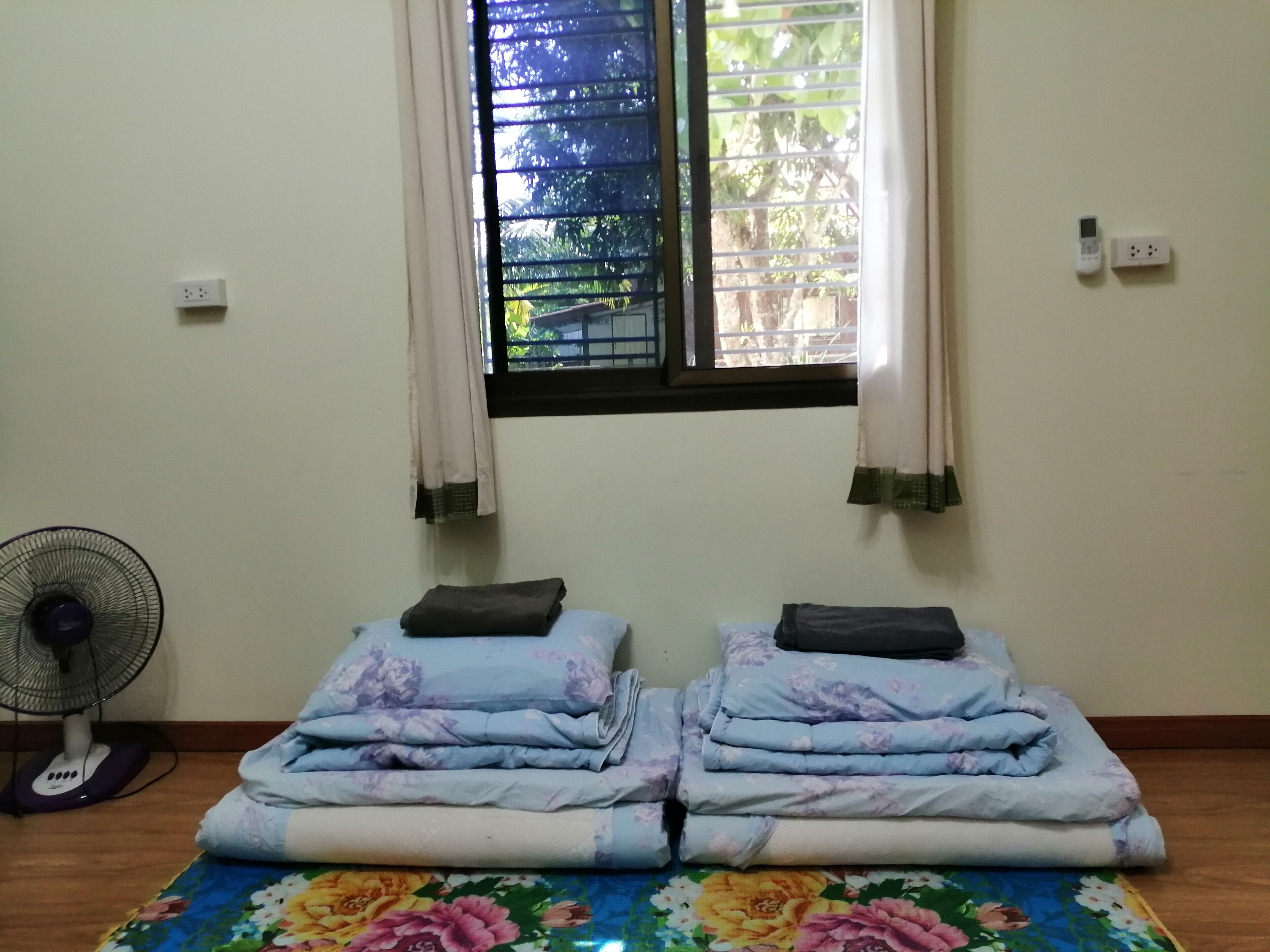 A room with two beds, a standing fan, and a window with curtains.