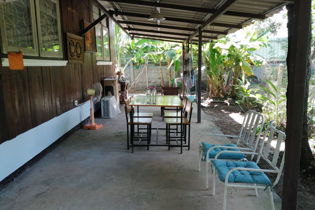 Um espaço coberto ao ar livre com mesa e cadeiras, cercado por plantas e um muro de madeira.