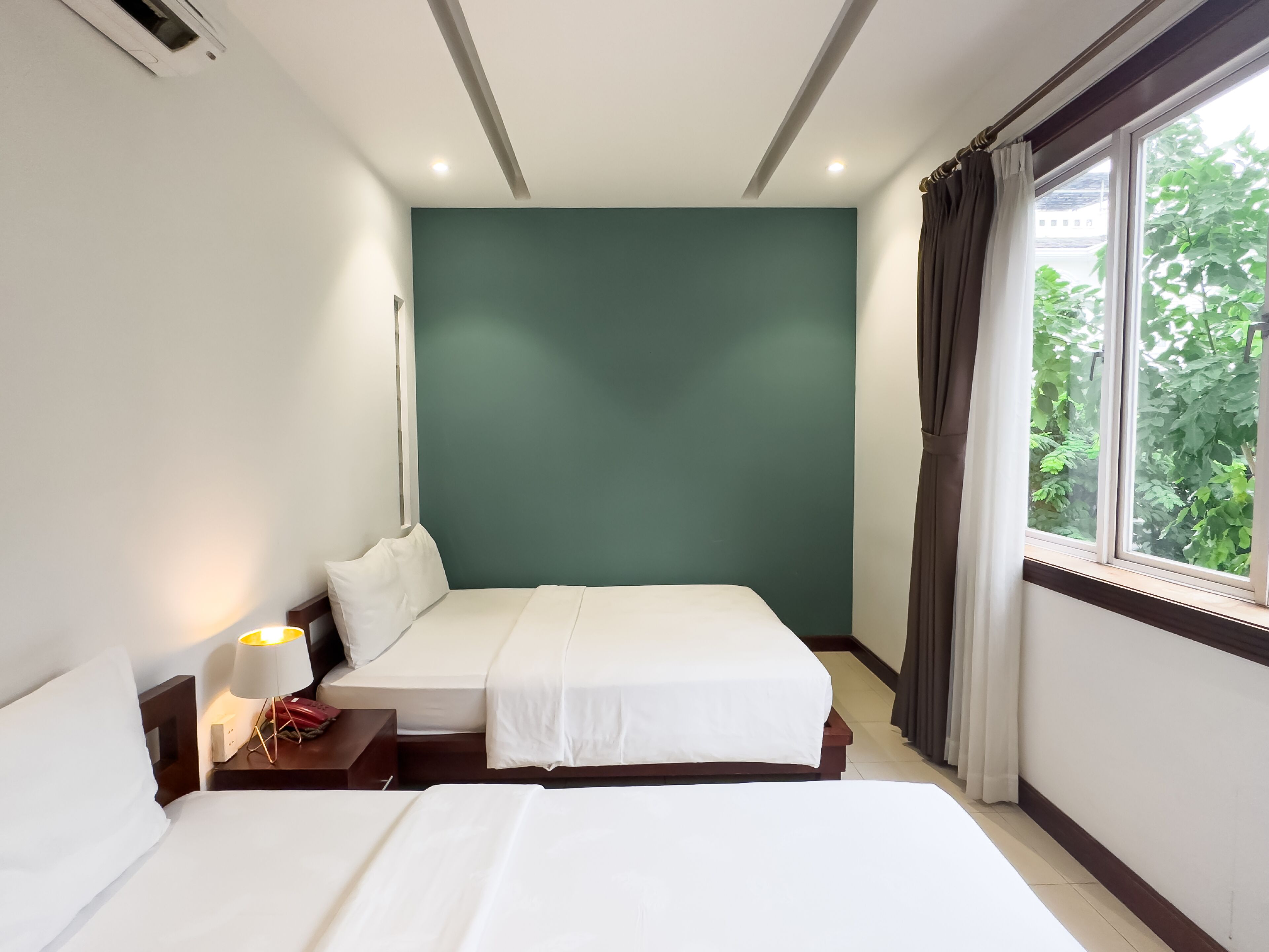 A hotel room with two beds, a nightstand with a lamp, a green accent wall, and a window with curtains.