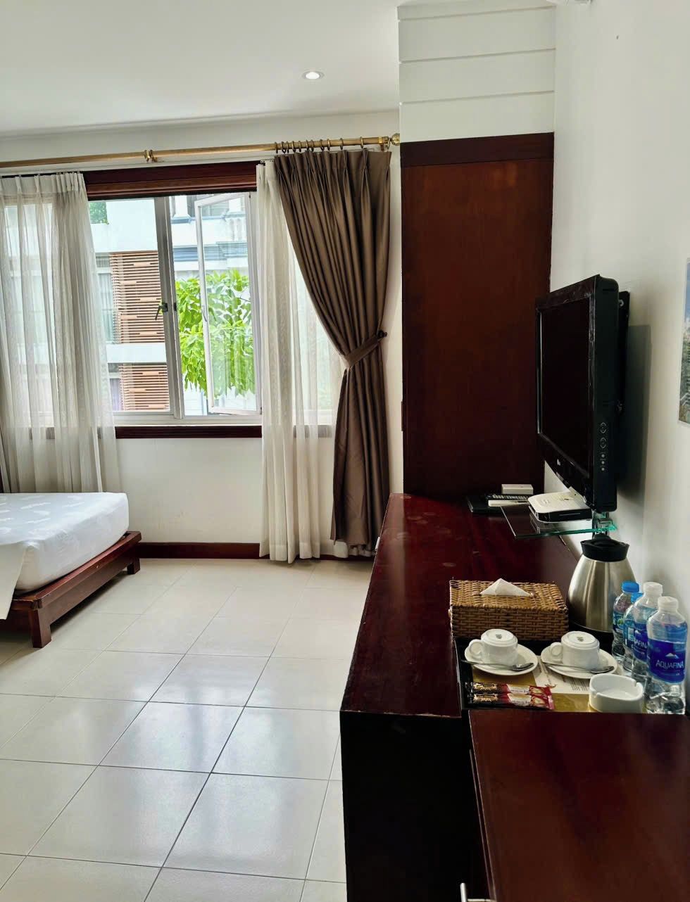 A hotel room with a bed, a TV, a wooden desk with a coffee setup, and a window with curtains.