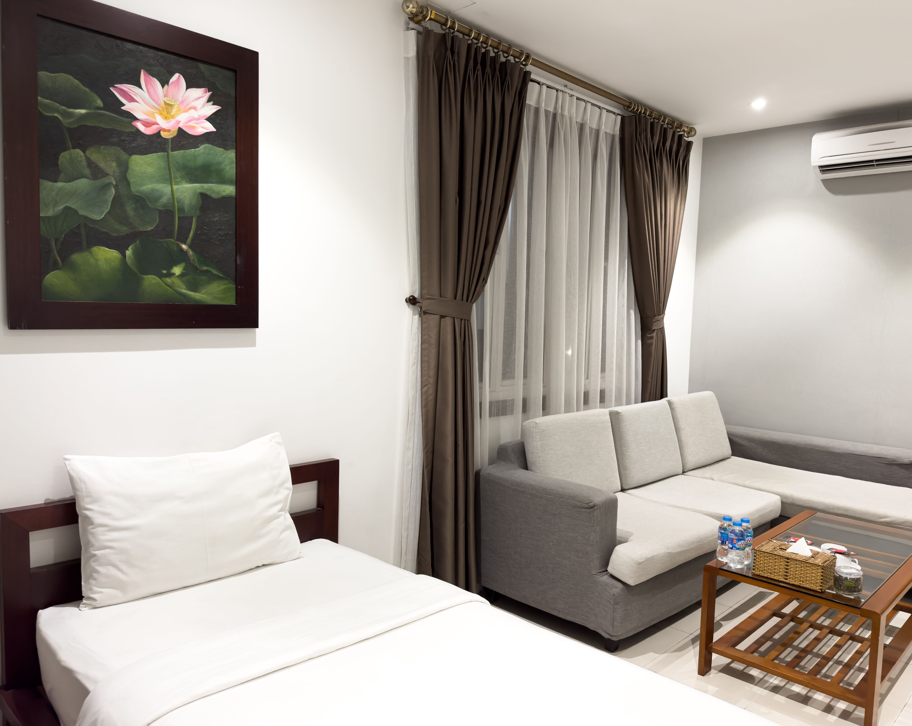 A modern hotel room with a bed, a sofa, a glass coffee table, and a painting of a lotus flower.