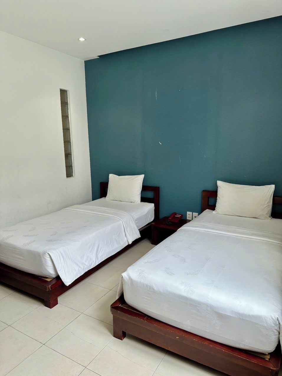 A hotel room with two single beds, a small bedside table with a lamp, and a window with blinds.