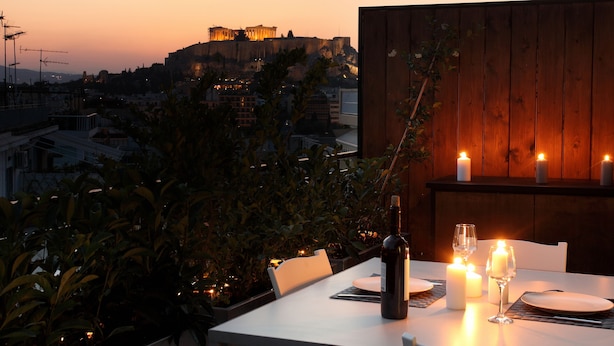 Mesa posta para dois, com velas, uma garrafa de vinho e vista para um edifício histórico ao pôr do sol.