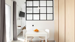 Uma sala de jantar moderna com uma mesa branca, duas cadeiras, uma tigela de frutas e uma televisão na parede.