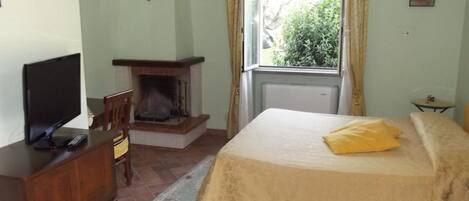 Quarto com cama, televisão em cómoda de madeira, lareira e janela com cortinas.