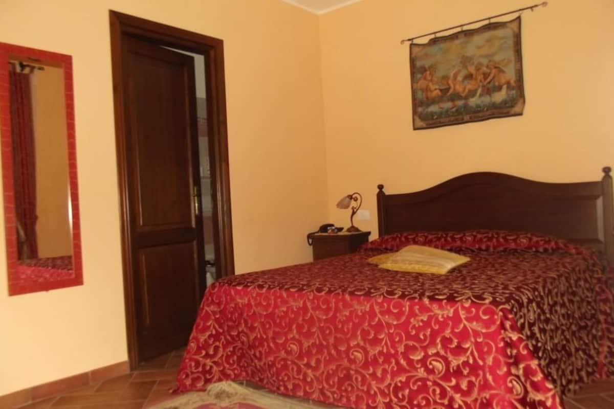 A bedroom with a wooden bed, a red patterned bedspread, a nightstand with a lamp, a mirror with a red frame, and a painting on the wall.