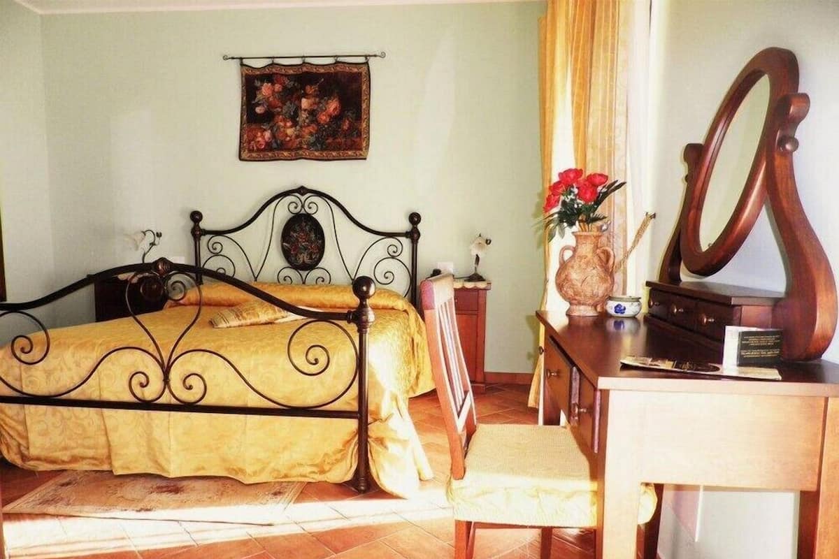 A bedroom with a bed, a desk, a chair, a mirror, and a vase with flowers.