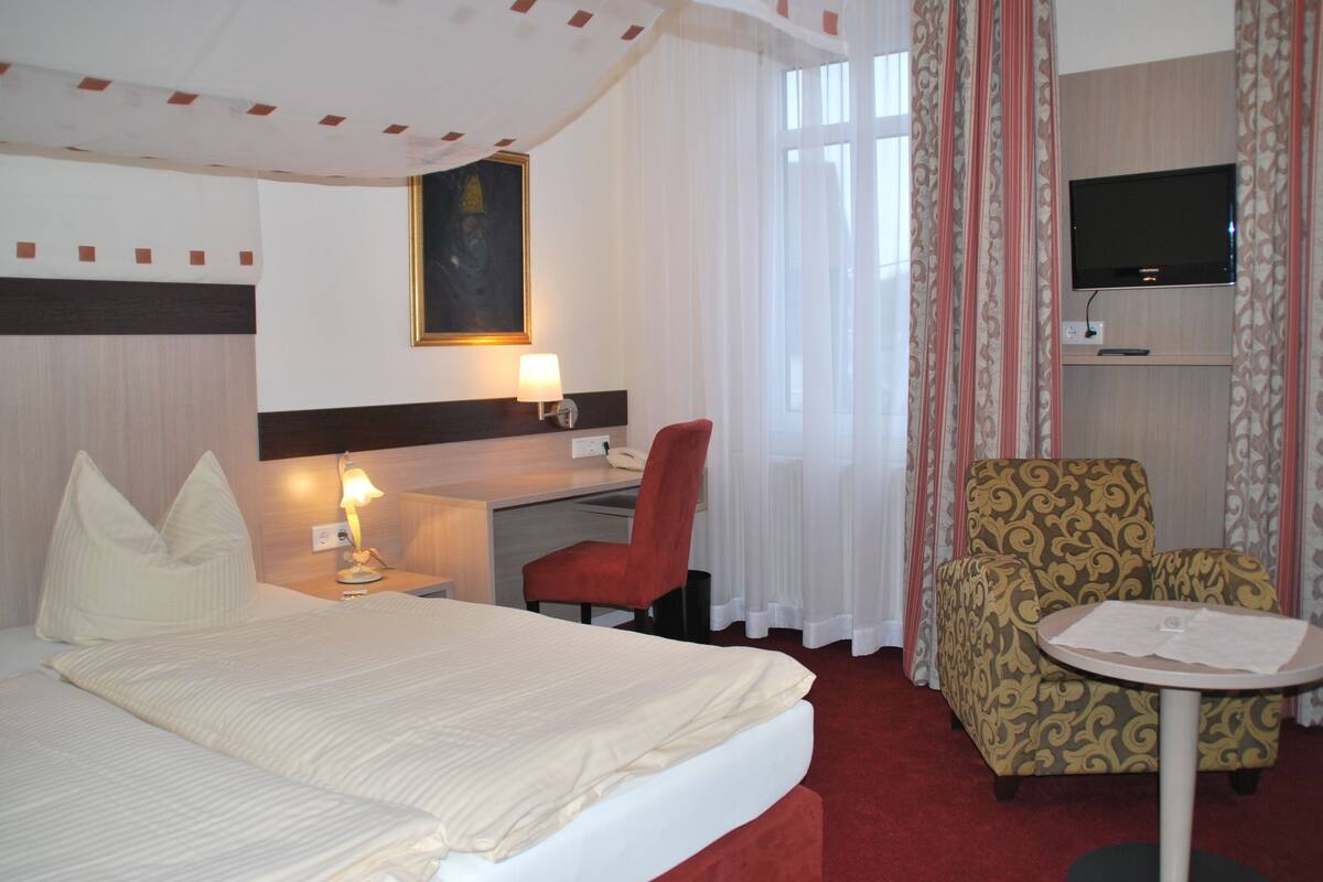 A hotel room with a bed, a desk with a lamp, a chair, a television on a stand, and a small table.