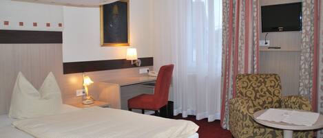A hotel room with a bed, a desk with a lamp, a chair, a television on a stand, and a small table.