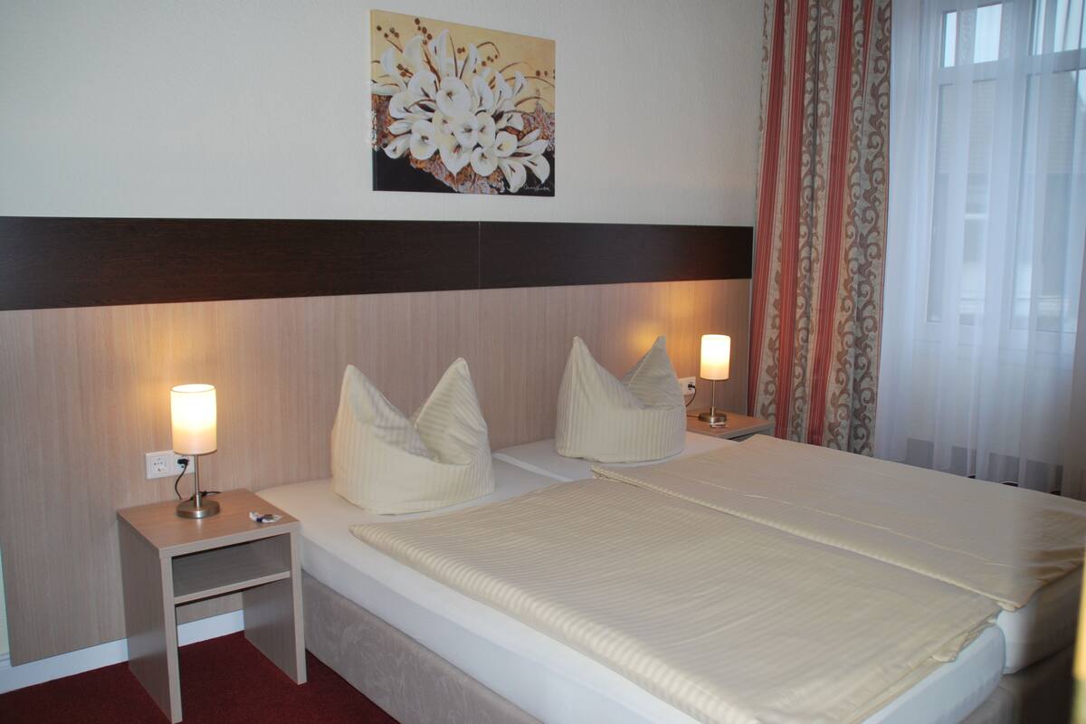 A hotel room with a bed, two bedside tables, lamps, a painting, and curtains.