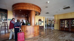 A reception area with a wooden reception desk, a red suitcase, and a person standing nearby. - Hotel ELE Puerta de Monfragüe (Malpartida de Plasencia)