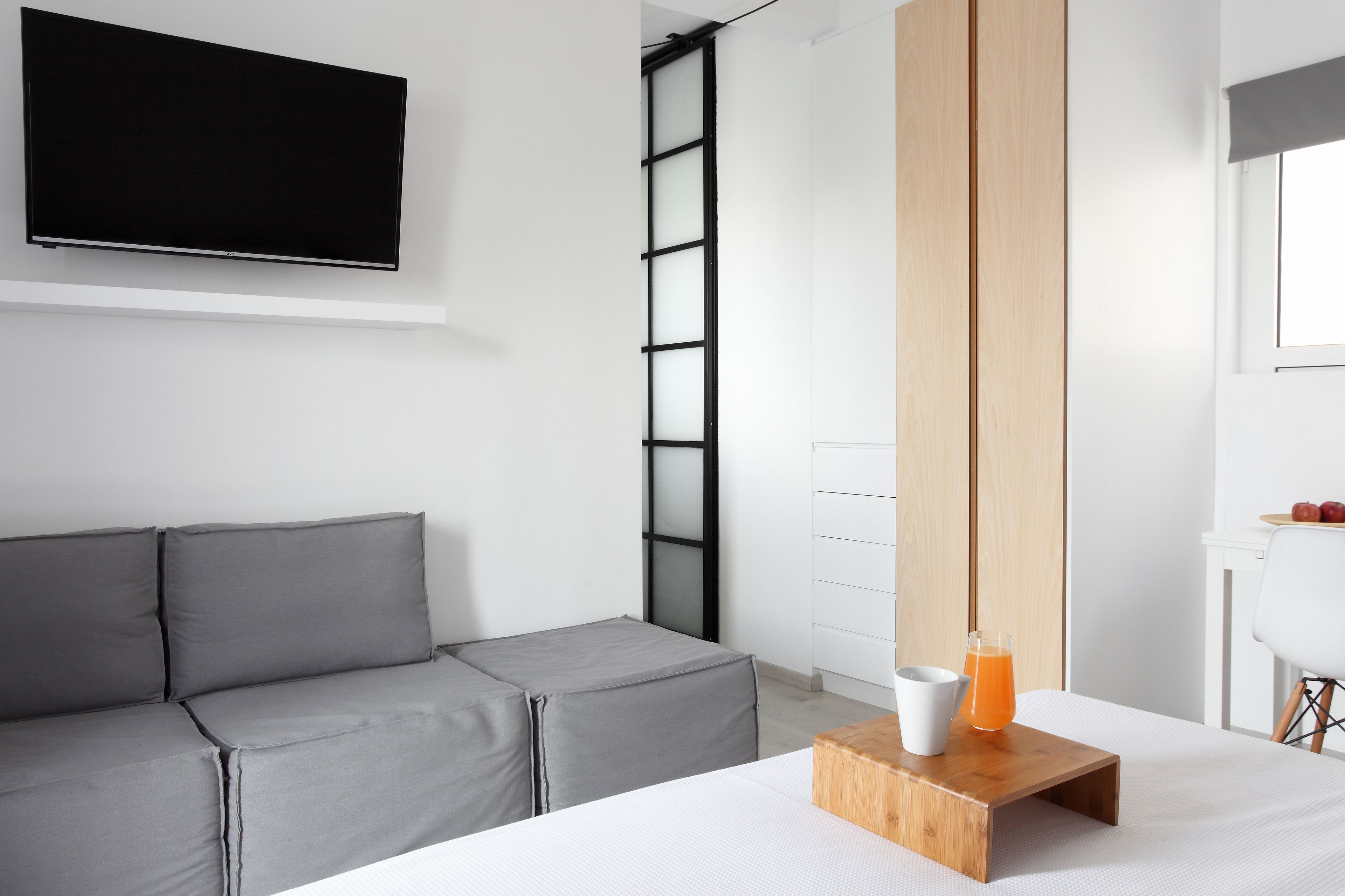 A modern hotel room with a grey sofa, a flat-screen TV mounted on the wall, a wooden bedside table with a glass of orange juice, and a white chair.