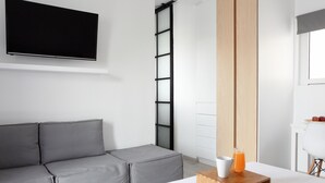 A modern hotel room with a grey sofa, a flat-screen TV mounted on the wall, a wooden bedside table with a glass of orange juice, and a white chair. - Athens View Loft - 04 (Athens)