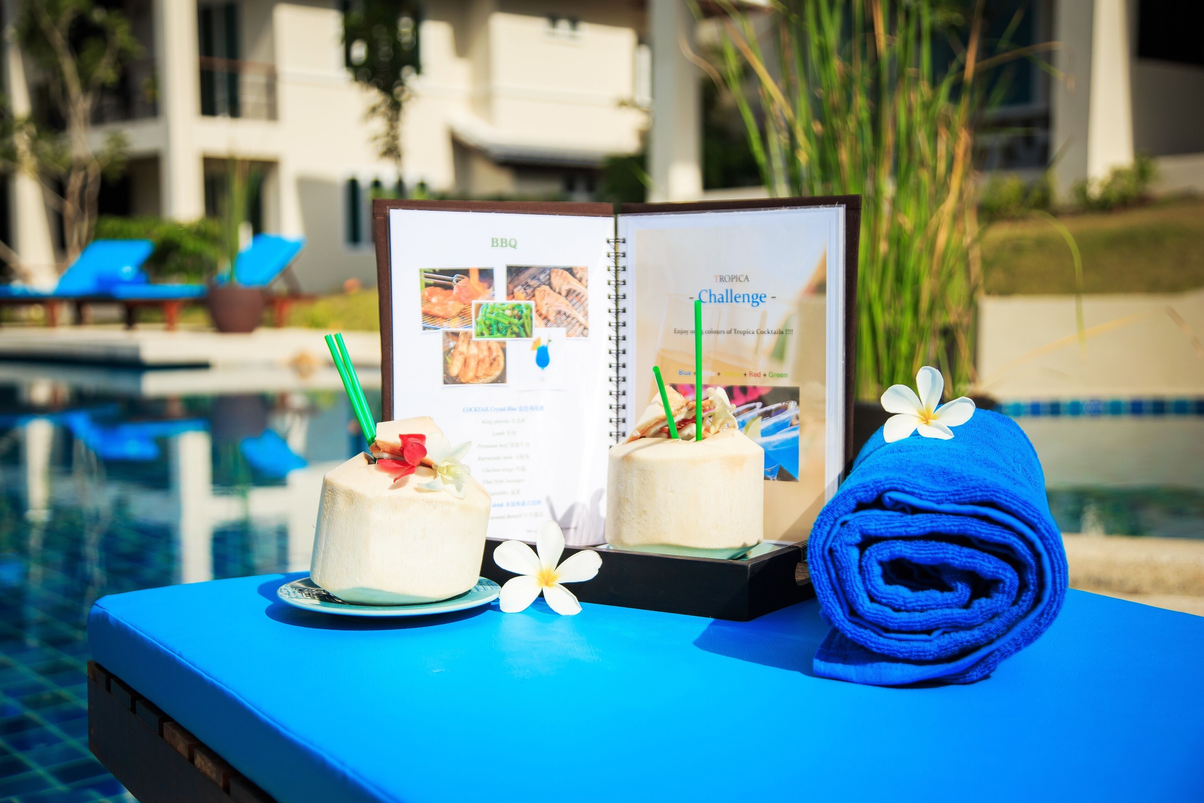 A poolside table with a menu, two cakes, and a rolled towel.