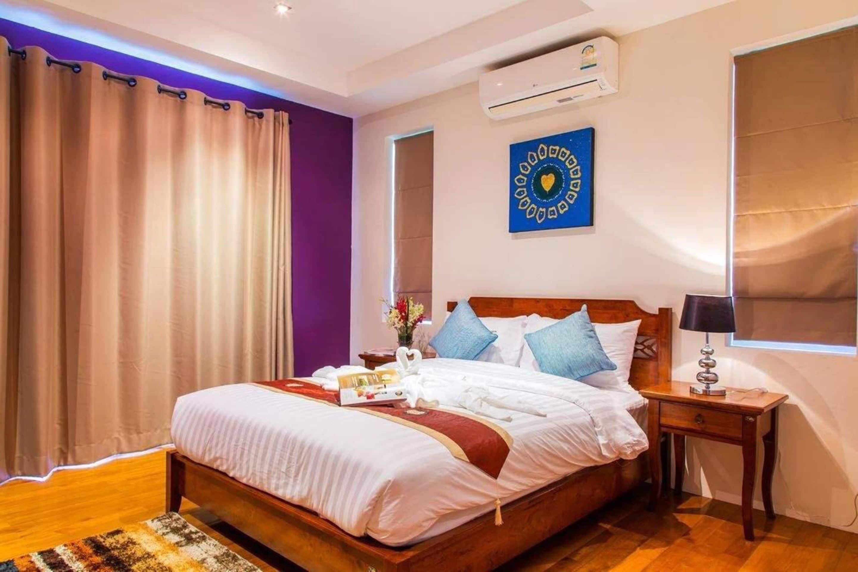 A neatly made bed with white linens and a red throw, a wooden nightstand with a lamp, and a wall-mounted air conditioner.
