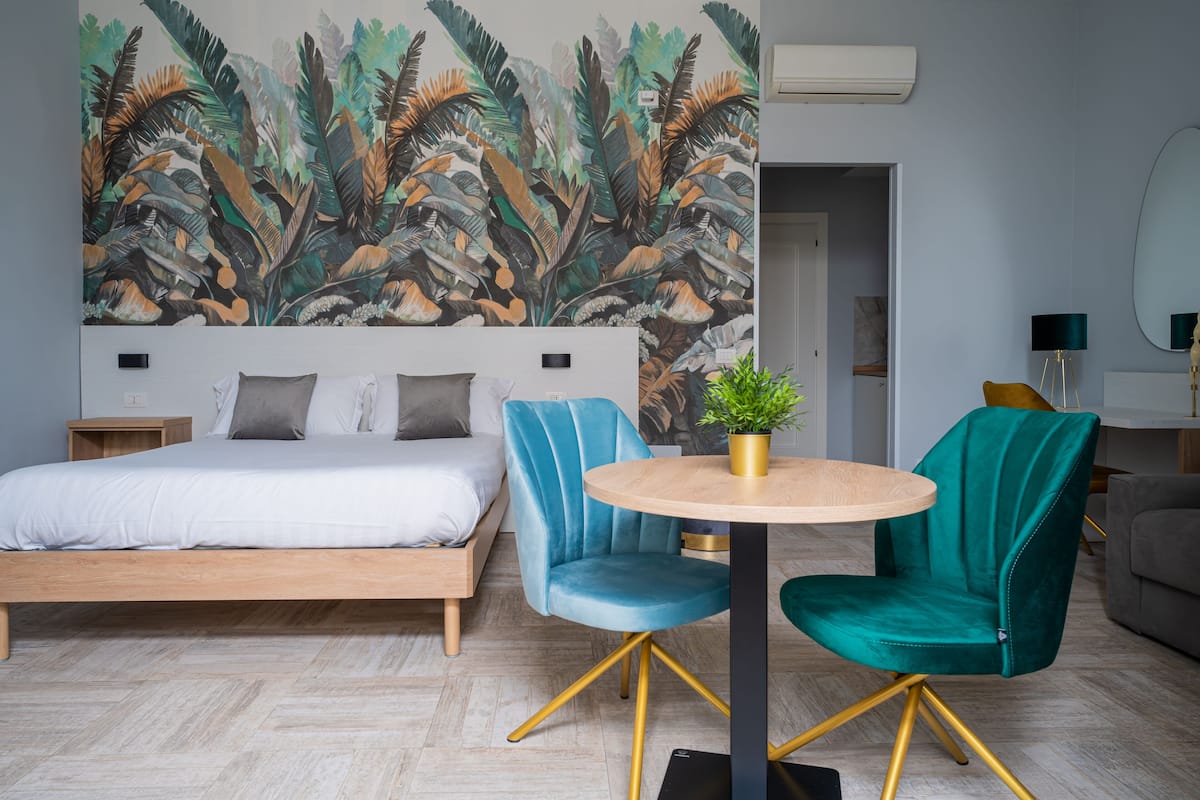 A modern hotel room with a large bed, two teal chairs, a small round table, and a tropical mural on the wall.