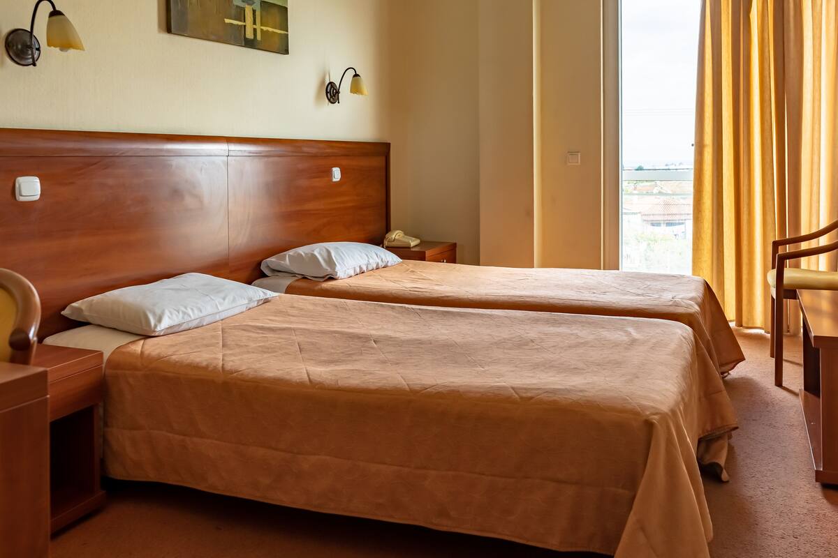 Une chambre d’hôtel avec deux lits simples, des têtes de lit en bois, des tables de chevet, une chaise et une fenêtre avec des rideaux.