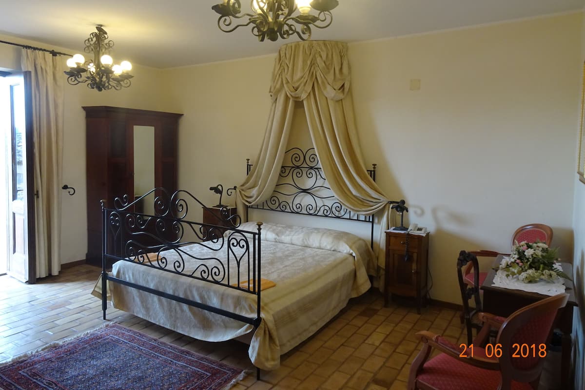 A bedroom with a bed, a chandelier, a wooden wardrobe, a chair, and a table with a vase.
