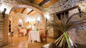A rustic dining area with stone walls, wooden beams, and a tiled floor. Tables are set with white tablecloths and red chairs. - Castello di Altomonte (Altomonte)