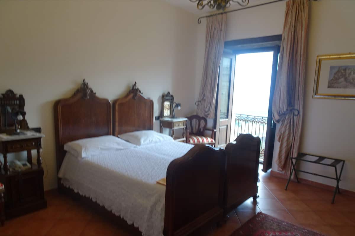 A bedroom with a wooden bed, a small bedside table, a chair, and a window with curtains.