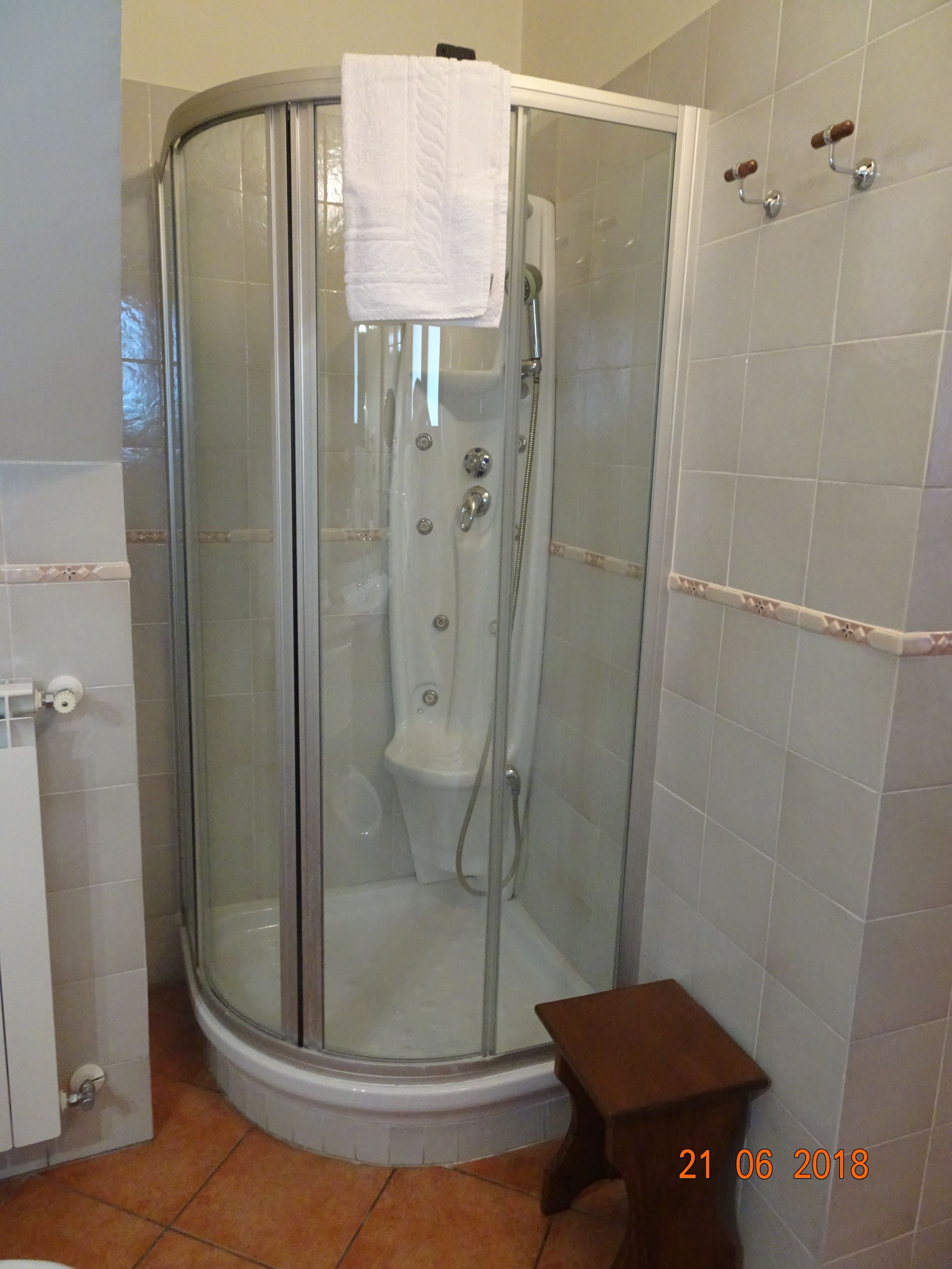 Cabina de ducha con paredes de vidrio, una toalla blanca colgada en la puerta, un cabezal de ducha manual y un pequeño taburete de madera.