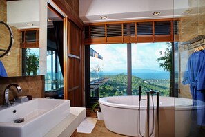 A modern bathroom with a large bathtub, a separate sink, and a large window offering a view of a mountainous landscape. - Luxury Villa Crystal Blue (Koh Samui)
