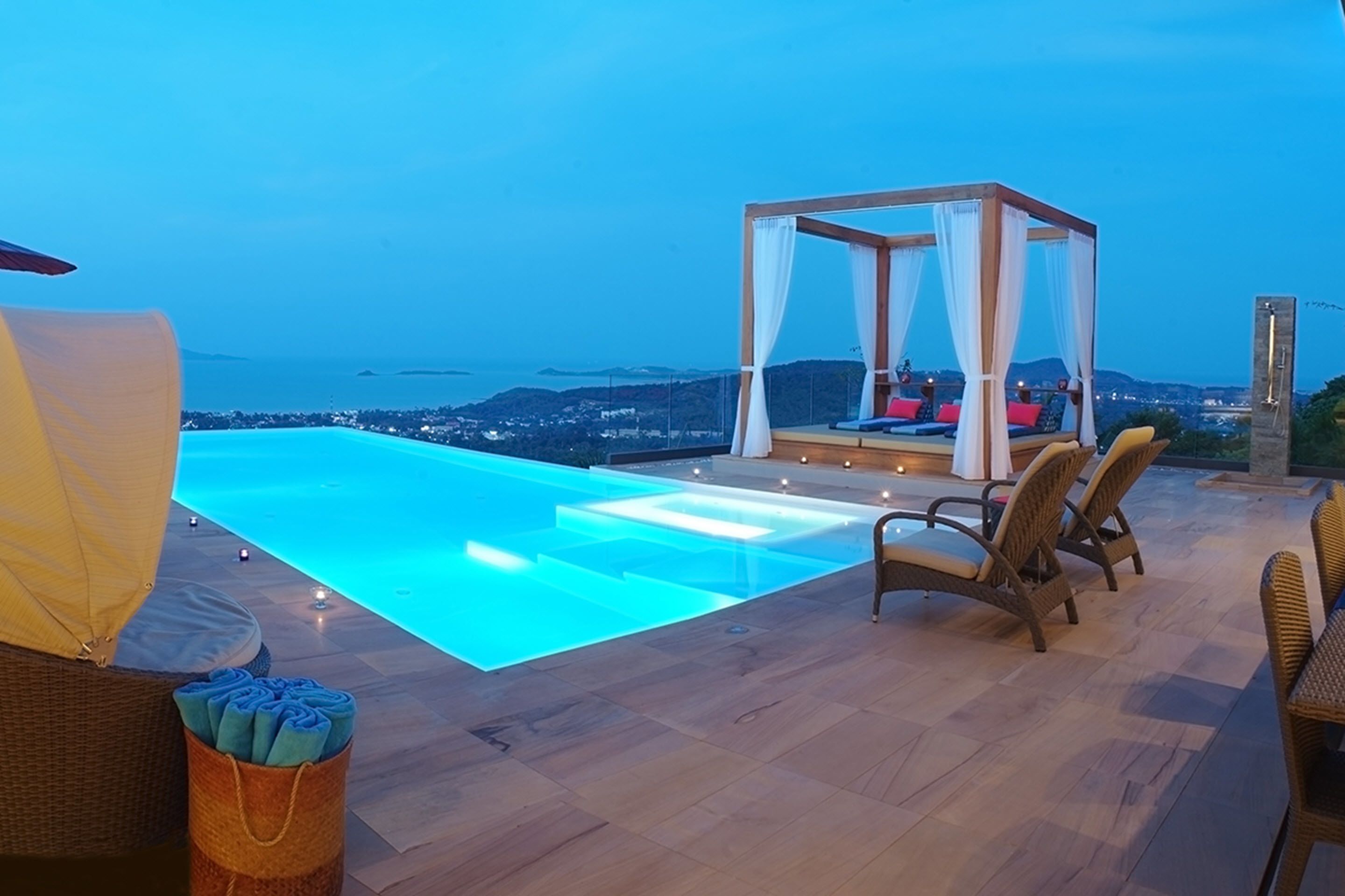 An infinity pool with a view of the city at dusk, a covered seating area with cushions, and wicker furniture.
