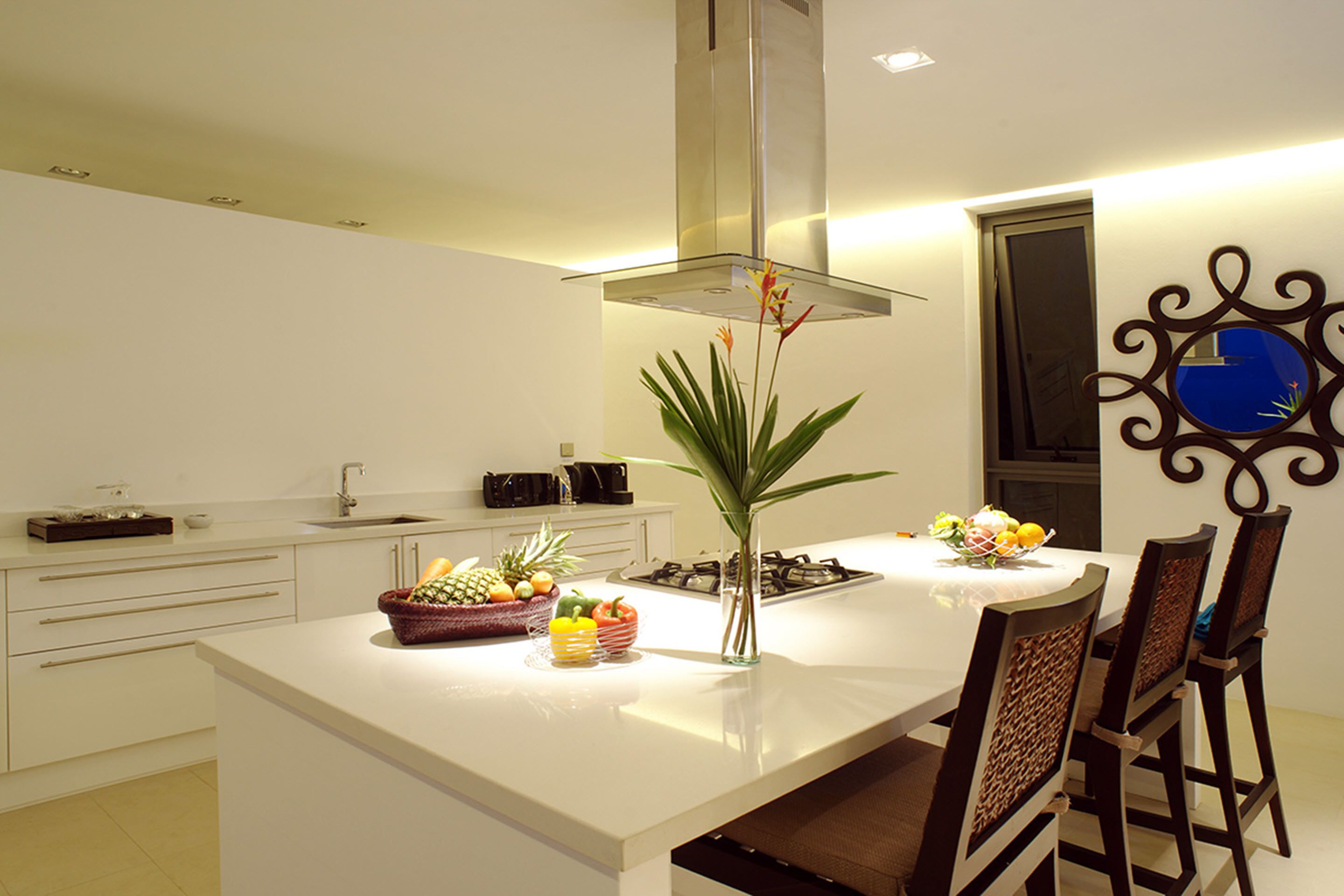 A modern kitchen with white cabinetry, a central island with fruit bowls, a stainless steel range hood, and a decorative wall mirror.