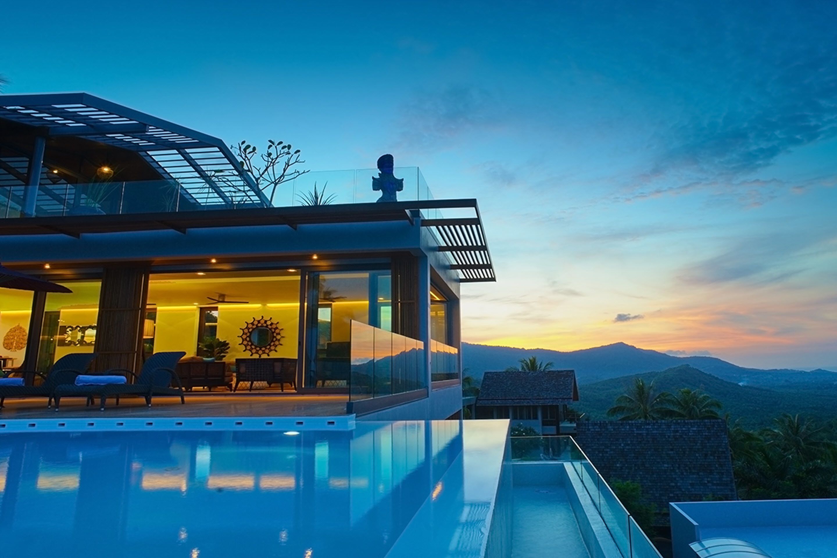 A modern house with a glass exterior, a pool, and a view of mountains at sunset.