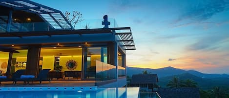 A modern house with a glass exterior, a pool, and a view of mountains at sunset.