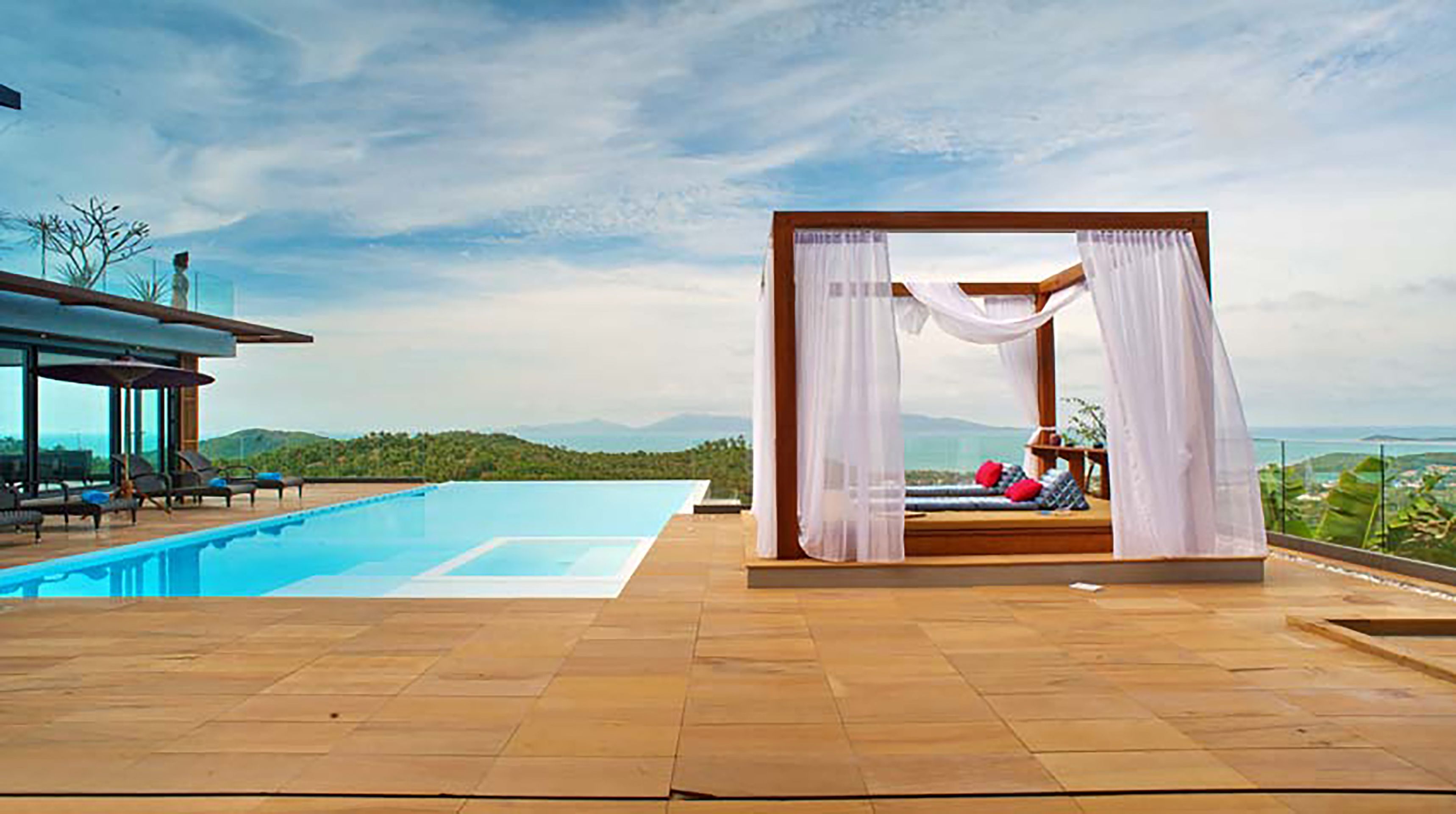 Zona de piscina con vistas despejadas a las montañas, un área de estar cubierta con cojines y un edificio moderno con piscina.