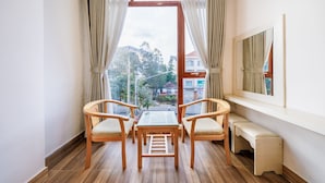 Een kamer met een houten vloer, een glazen tafel, twee stoelen, een bankje en een groot raam met gordijnen.