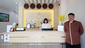 Een hotel receptiebalie met een receptioniste, een man die ernaast staat, en een muur met namen van steden.