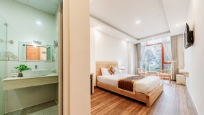 Een moderne hotelkamer met een houten bed, een groot raam met gordijnen, een badkamer met wastafel en spiegel, en een klein zithoekje.