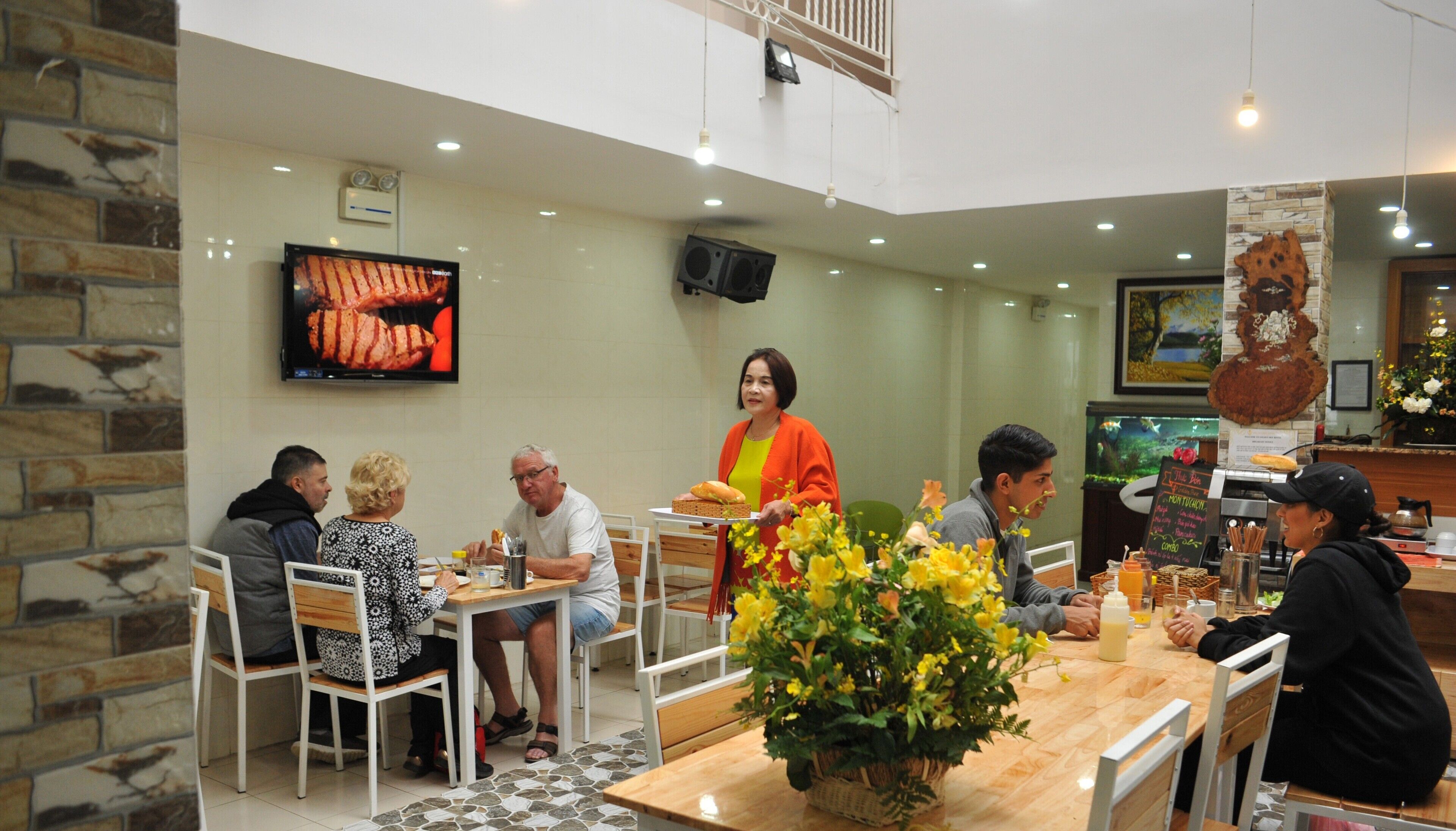 Un restaurant avec un mur en pierre, un écran de télévision diffusant des images de plats et un arrangement floral en guise de décoration de table.