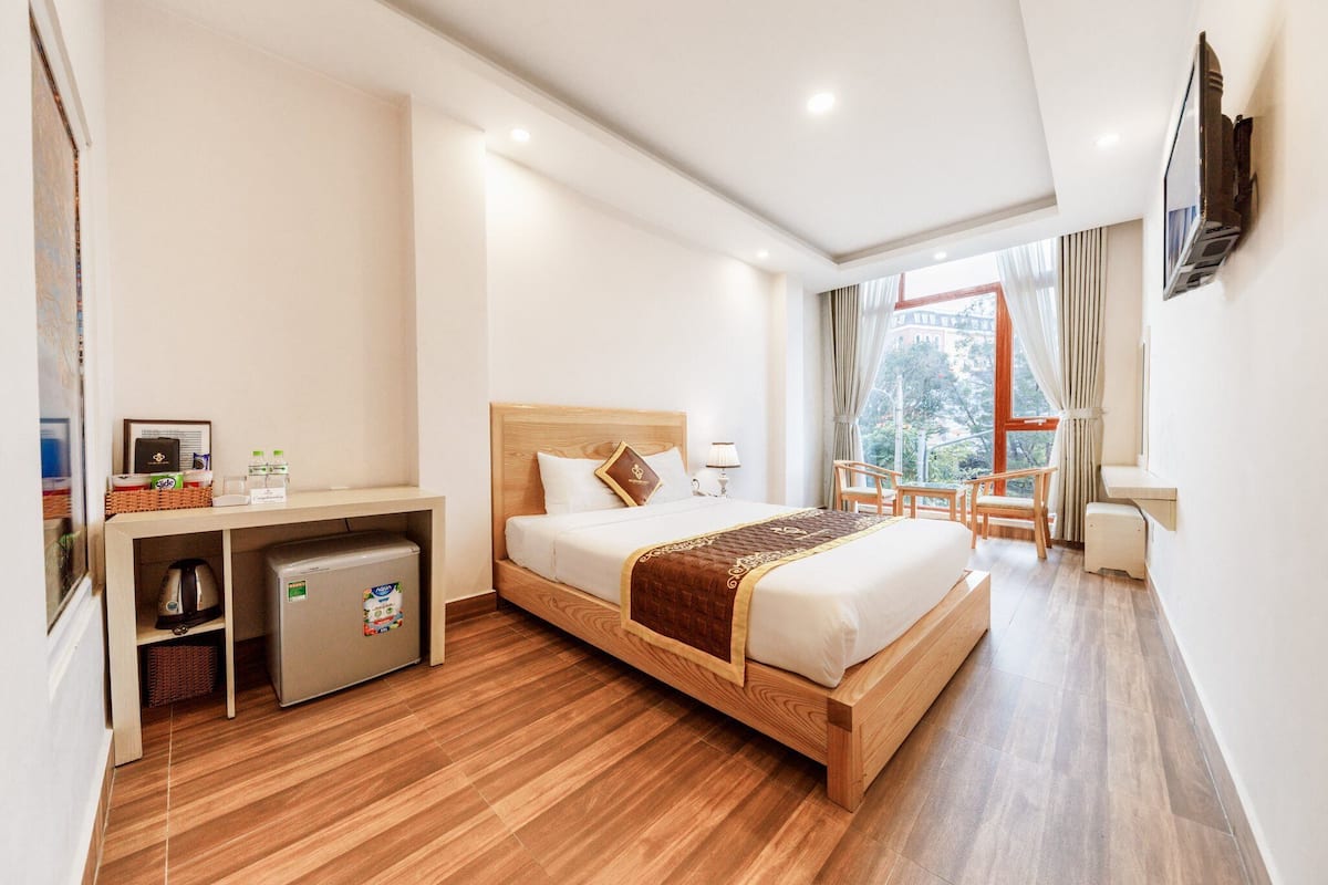 A hotel room with a wooden bed, a small refrigerator, a desk with a chair, and a window with a view of the outside.