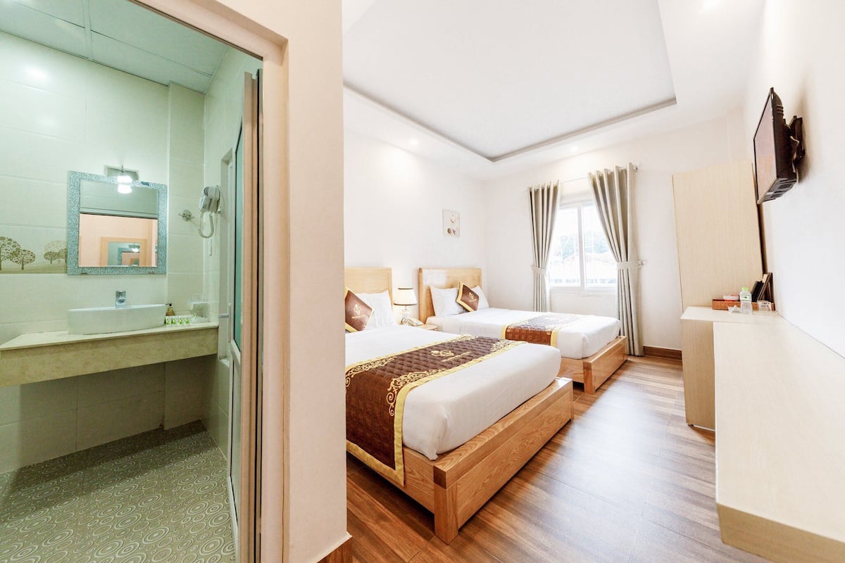 A hotel room with two beds, a wooden floor, a bathroom with a sink and mirror, and a window with curtains.
