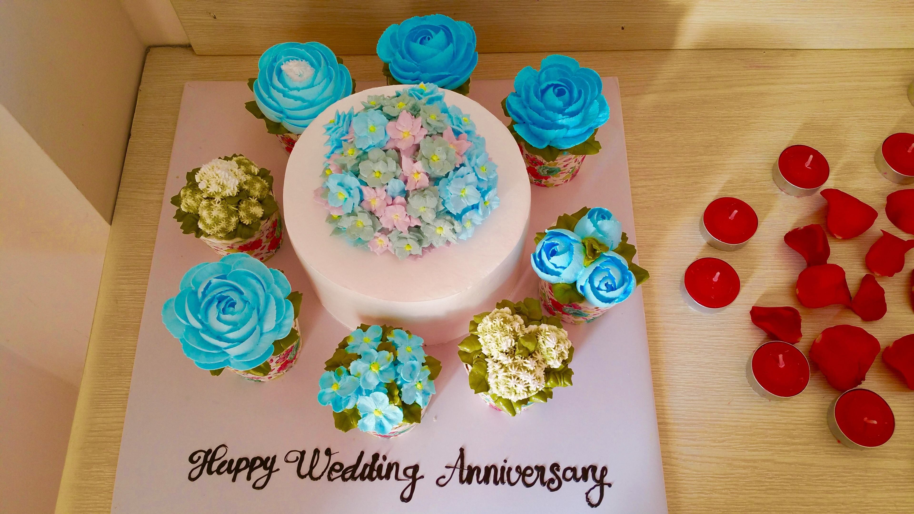 Taart met blauwe en roze bloemen, omringd door cupcakes en kaarsen op een kaartje met de tekst 'Happy Wedding Anniversary'.